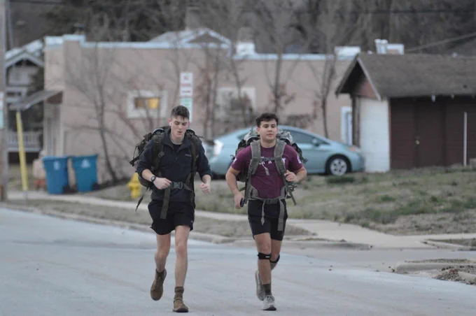 Two people are jogging on a suburban street wearing backpacks and casual outdoor clothing. Houses and cars are visible in the background.