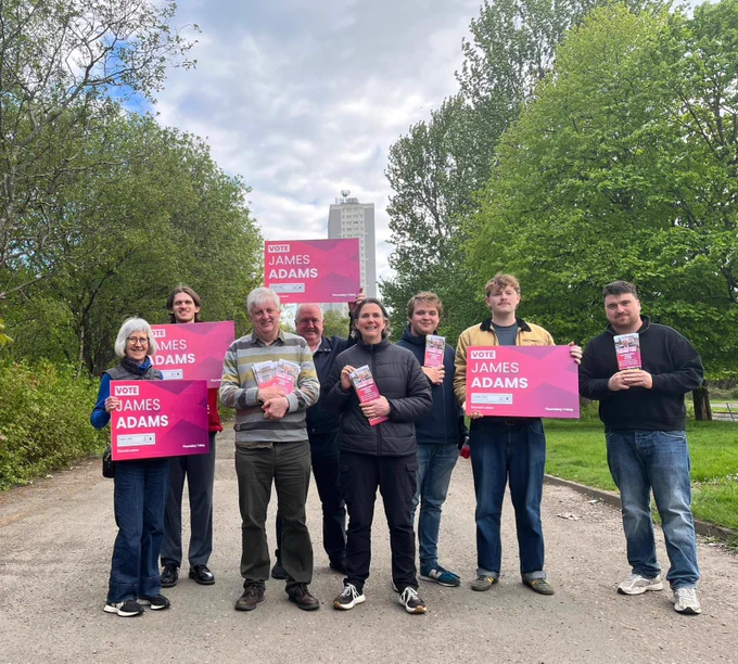 Candidate James Adams with Labour campaigners holding placards and leaflets, with trees and tower blocks in the background