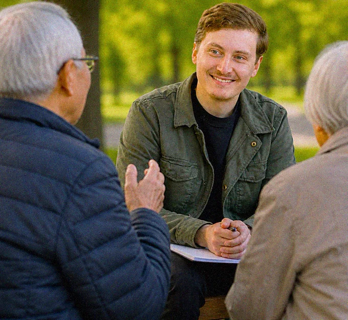 A man in a green coat, talking to a pair of seniors