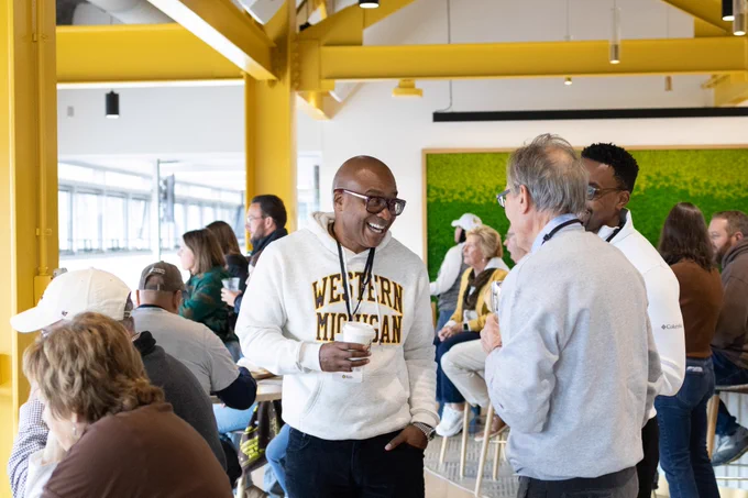 A group of people socializing in the Waldo
Stadium President's Suite. One person wearing a "Western Michigan" sweater is smiling while holding a coffee cup and talking to another person facing them. The room has bright yellow beams, and attendees are casually dressed, engaging in conversation.