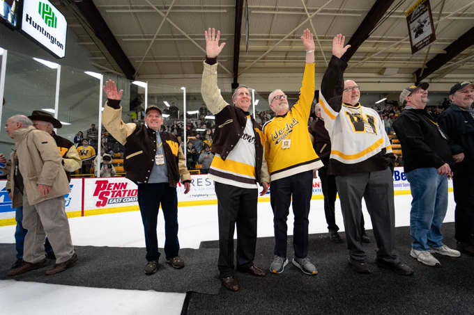 A group of alumni stand on the ice at Lawson Arena, wearing Western Michigan University apparel. They are waving and smiling at an audience. Banners and advertisements line the rink's boards, and spectators are visible in the stands.