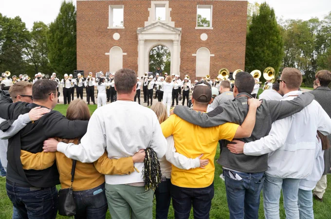 A group of alumni stand with their arms around each other, facing the Bronco Marching Band performing on the lawn of Heritage Hall. The band members are in uniform with brass instruments.