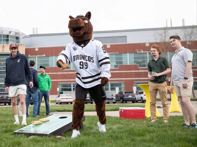 Buster Bronco playing cornhole on a grassy area. Several people stand around watching, and a few buildings are in the background.