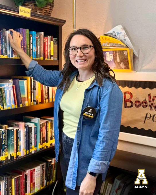 Jeana Link ’09 stands in a classroom library, smiling and reaching for a bookshelf while wearing a “Forever a Mountaineer” button on her shirt.