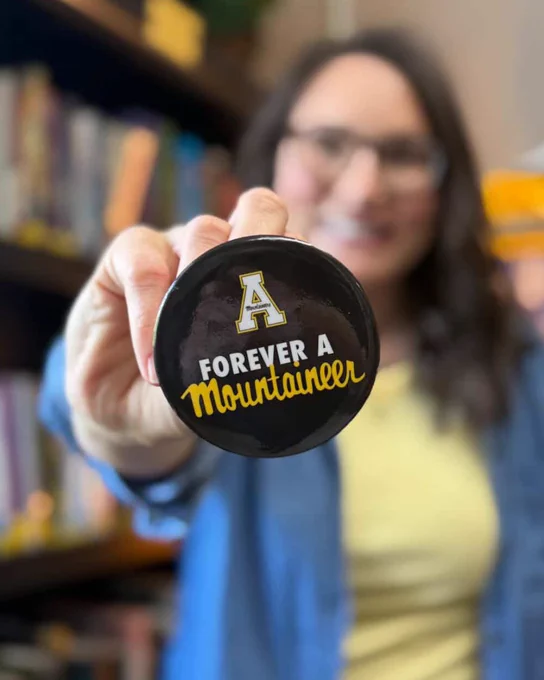 Close-up of Jeana Link ’09 holding a “Forever a Mountaineer” button toward the camera, with a classroom bookshelf blurred in the background.