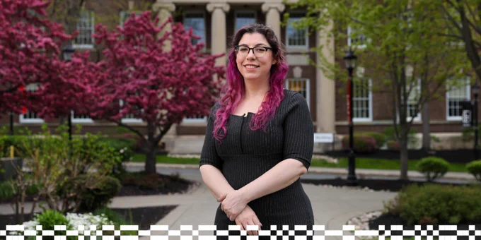 Portrait of a person with long wavy dark hair dyed pink at the ends, wearing glasses and a dark pinstriped dress, standing with arms crossed in front of a columned campus building. A blooming tree with bright pink flowers and green foliage frames the background.