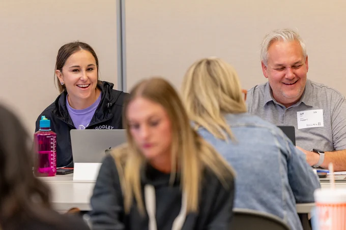 A group of educators is sitting at a table, working on laptops. One person has a pink water bottle, and there are name tags visible. 