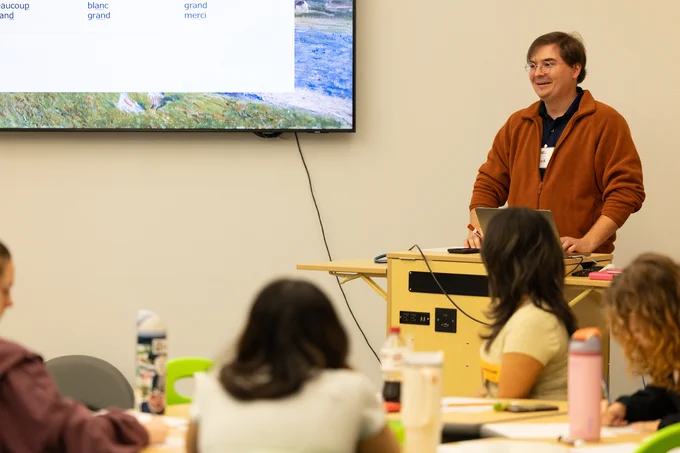 A GoTeach11 candidate is presenting at the front of a room, standing behind a podium with a laptop. The presentation is displayed on a screen featuring a list of French words. Seated students are facing the presenter, taking notes.
