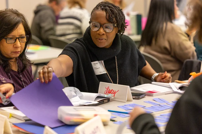 A person wearing glasses, seated at a table with others, is engaged in sorting colorful papers and name cards on the table. 