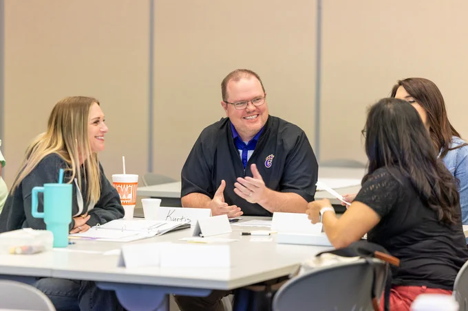 A group of people sit around a table engaged in conversation. Papers, name tags, and drinks are on the table. One person is smiling and gesturing while others listen attentively.