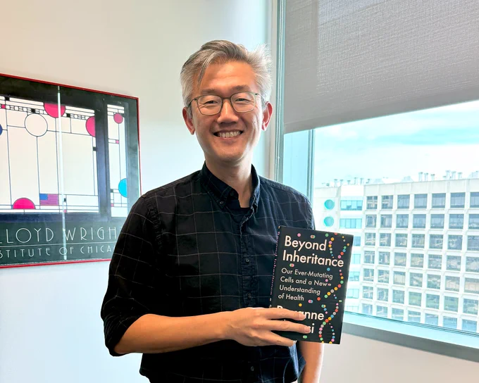 Hao Zhu stands in front of a window holding the book "Beyond Inheritance: Our Ever-Mutating Cells and a New Understanding of Health."