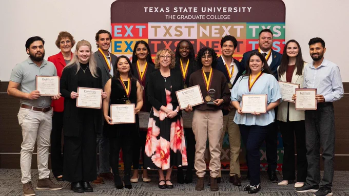 Members of the Texas State University graduate house of representatives holding certificates and wearing medals in front of a university banner.