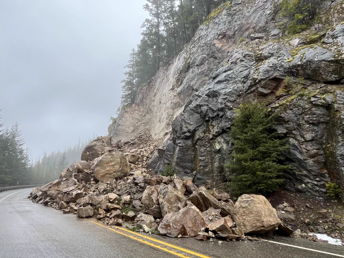 A rockslide with the slope on the right and a pile of rocks, boulders and debris at the base cover State Route 20 beyond the center line of the highway on a cloudy day.