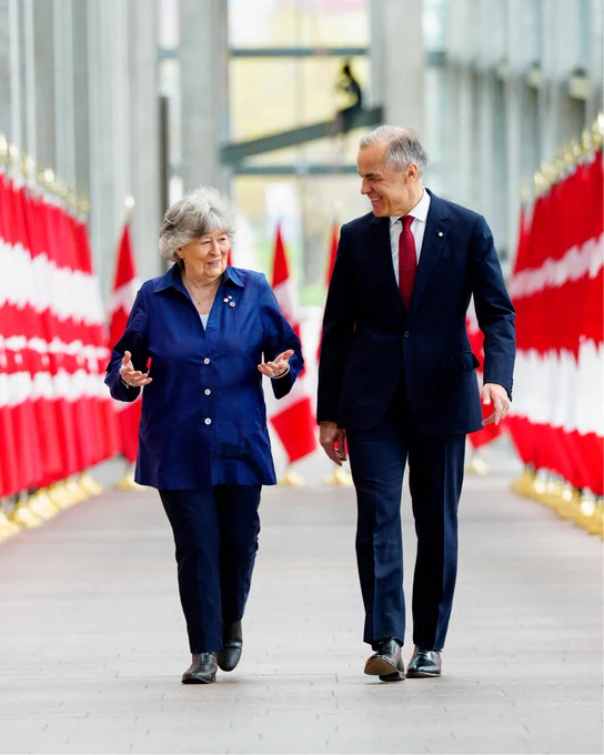 Prime Minister Mark Carney walks alongside the Honourable Louise Arbour, speaking together in a corridor lined with Canadian flags.