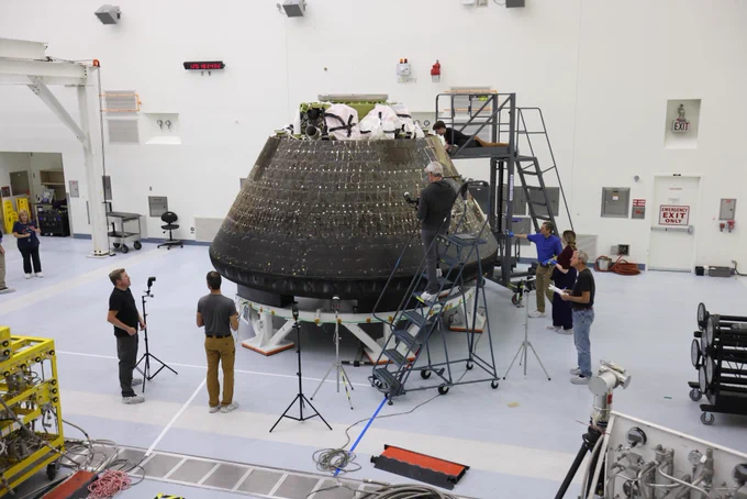 The Artemis II Orion crew module sits on a stand inside the Multi-Payload Processing Facility at NASA\u2019s Kennedy Space Center. Several technicians stand on mobile platforms and ladders around the spacecraft, inspecting its darkened exterior. Additional personnel stand nearby with tripods and equipment. The room contains tools, hardware, and machinery arranged around the perimeter, with white walls and a high ceiling. Photo credit: NASA/Ben Smegelsky