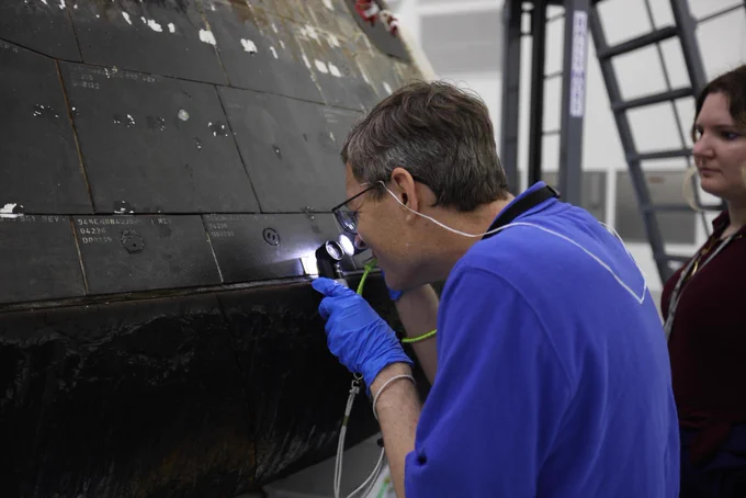 A close-up view of NASA\u2019s Orion spacecraft from the Artemis II mission shows a technician carefully inspecting the capsule\u2019s darkened backshell tiles with a lighted magnifier. Another team member stands nearby as the spacecraft undergoes de-servicing inside Kennedy Space Center\u2019s Multi-Payload Processing Facility. Photo credit: NASA/Ben Smegelsky