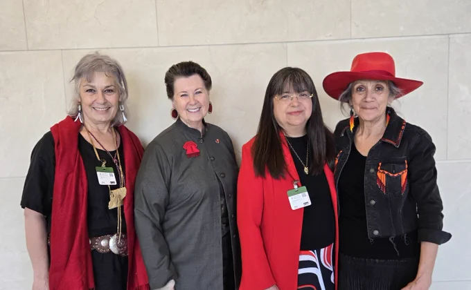 Four women stand together, smiling at the camera, against a white wall. 

The women are wearing various items of red to represent Red Dress Day. 