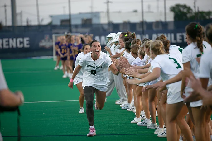 Sofia Chepenik, wearing a USF jersey, runs through a line of cheering teammates on a sports field, high-fiving them. The background shows another team in purple and yellow uniforms.