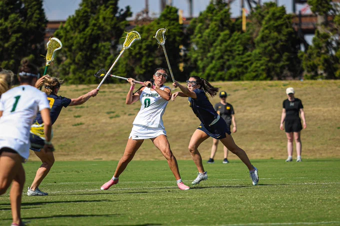 A lacrosse game in progress on a field. Sofia Chepenik, wearing a white uniform labeled number 0, is holding a lacrosse stick and moving towards the goal. They are closely pursued by a player in a dark kit. Additional players are visible in the background.