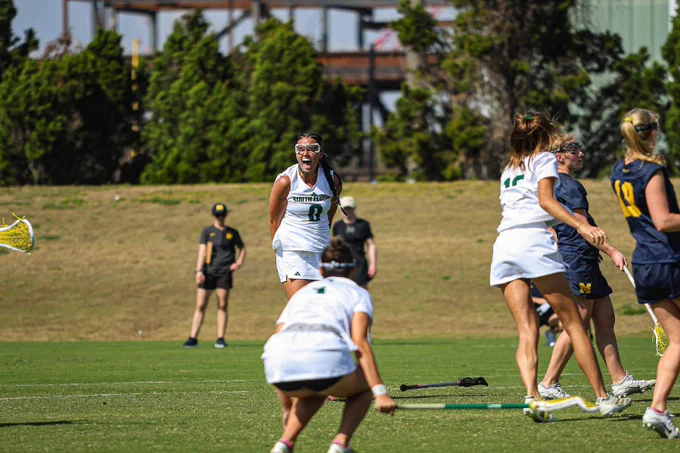A lacrosse game in progress on a field. Sofia Chepenik, wearing a green and white uniform, is energetically reacting, holding a lacrosse stick. Other players are engaged in the game. A coach wearing a dark outfit watches from the sideline.