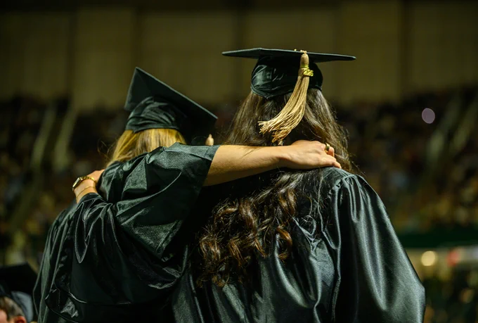 Graduates in black caps and gowns stand with their arms around each other, facing a blurred audience.