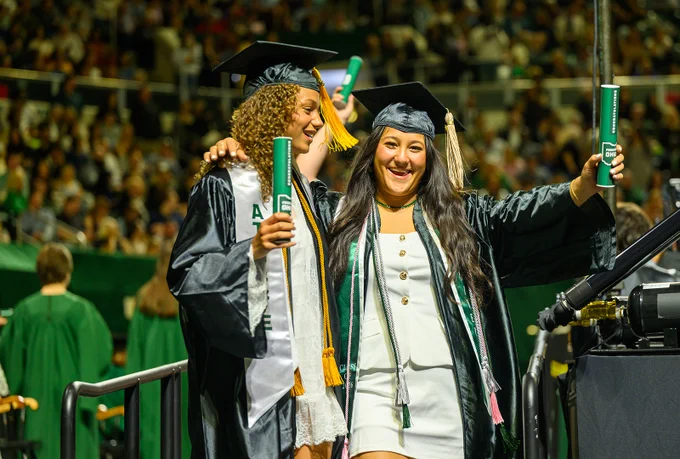Two graduates in academic caps and gowns celebrate. A crowd of people is visible in the background.