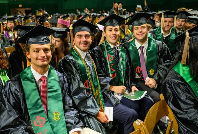 A group of graduates sitting together, wearing black gowns and caps with green stoles featuring Greek letters. They are smiling and holding programs, surrounded by a large audience in similar attire at a graduation ceremony.