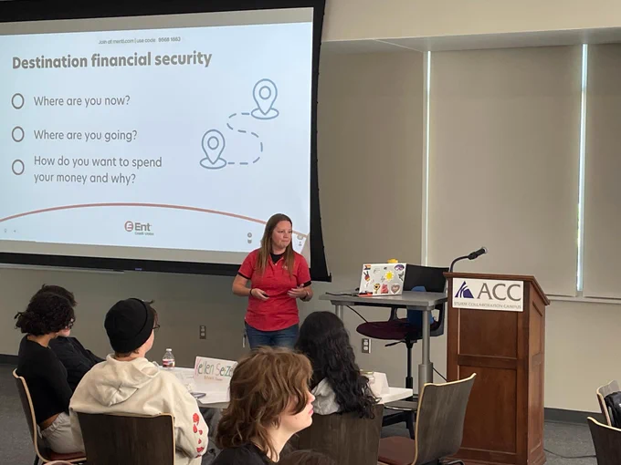 A person presenting in a classroom with a slide titled "Destination financial security," showing three bullet points: "Where are you now?", "Where are you going?", and "How do you want to spend your money and why?". The Ent Credit Union logo is visible on the slide. The room has seated individuals and a podium with the ACC logo.