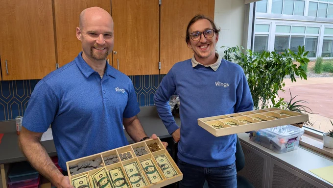 Two people are smiling and holding trays filled with cash and coins. They are standing in an office setting with indoor plants and large windows visible in the background. Both are wearing shirts with the "Wings" logo.