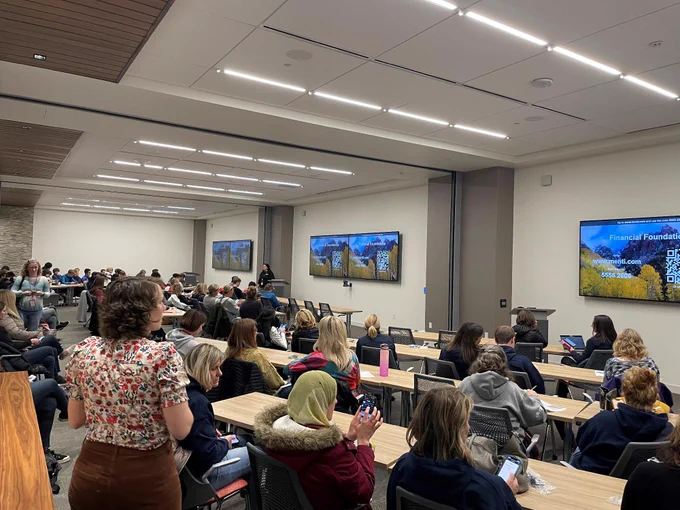 A large classroom filled with attendees seated at tables facing forward. Multiple screens display a presentation titled "Financial Foundations" with a QR code and contact information. The room has bright lighting and modern design.