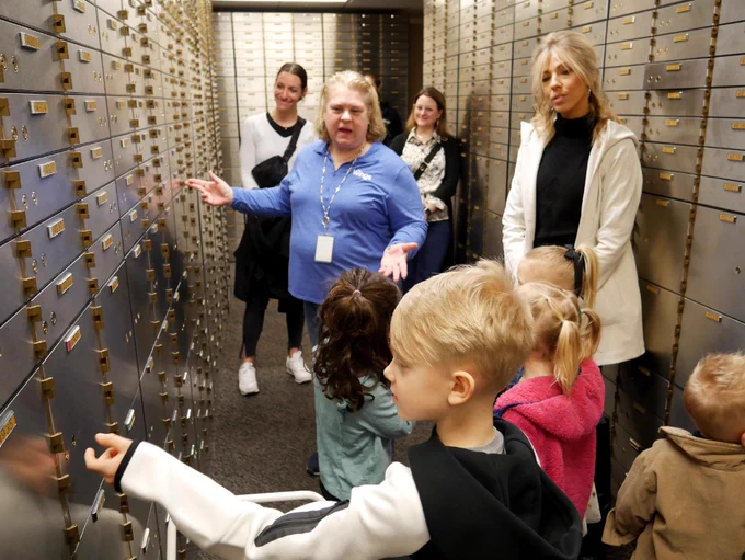 A group of people, including adults and children, are standing in a narrow hallway lined with safety deposit boxes. An adult is gesturing towards the boxes while others observe. The setting appears to be a bank vault.