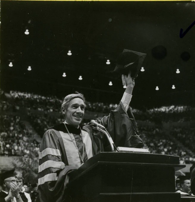 Black-and-white photograph of a person in academic regalia standing at a podium in a large arena, raising a graduation cap overhead while speaking into a microphone, with a seated audience visible in the background under overhead lights.