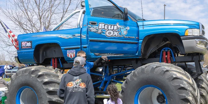 A large blue monster truck labeled "Blue Ox" is displayed on grass. Several individuals are gathered around it.