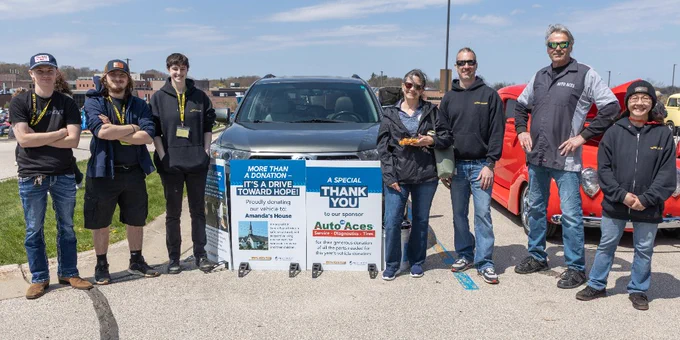 A group of people stands in front of parked cars in an open area.