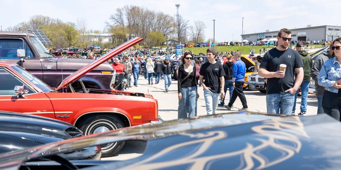 A lively car show scene with numerous attendees walking between classic cars.