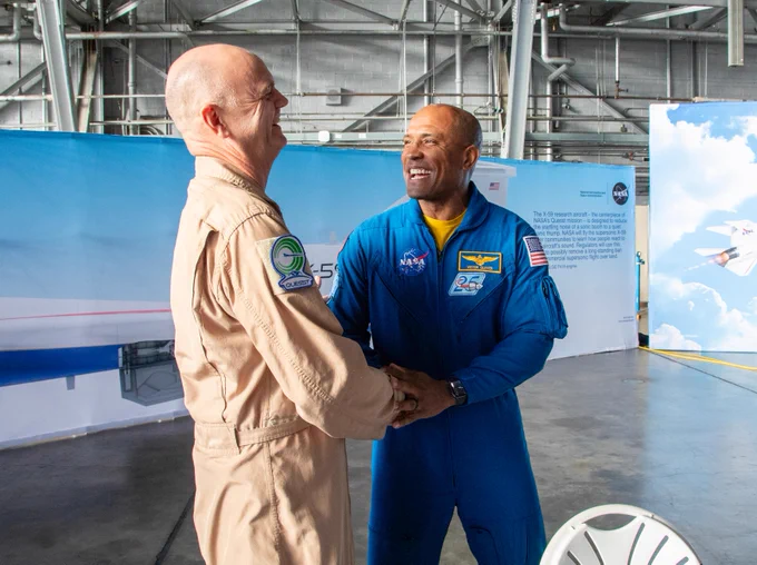 NASA astronaut Victor Glover (right), wearing a blue flight suit, smiles as he shakes hands with NASA test pilot Nils Larson (left) in a hangar at NASA\u2019s Langley Research Center. The two stand in front of an X-59 aircraft display, reconnecting years after Larson trained Glover as a flight instructor.