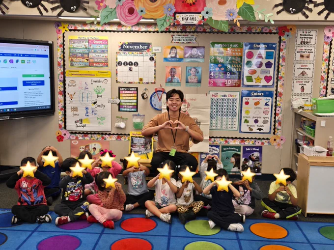 Henry Ngo in a classroom with his students, making heart shapes with their hands. The children's faces are covered with stars.