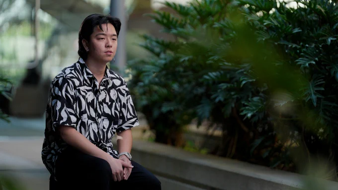 Henry Ngo sitting and looking away from the camera to the right. Foliage in the background.