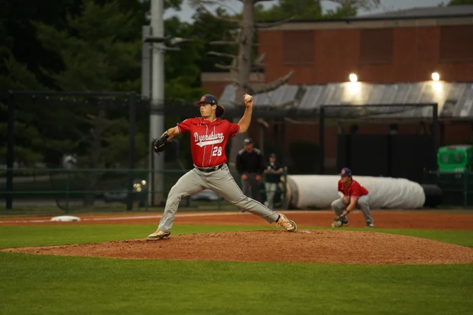 Baseball pitcher in a red jersey and gray pants winding up to throw a pitch from the mound during a game, with an infielder crouched and ready in the background on a well-lit field.
