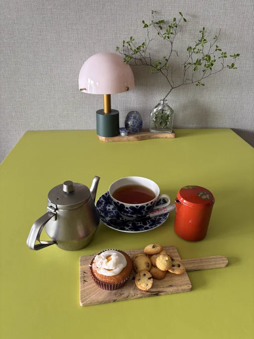 黄緑色のテーブルにステンレスのポット、青い花柄のカップに入った紅茶、赤い茶筒、そしてマフィンとクッキーが載った木製のプレートが並んでいます。