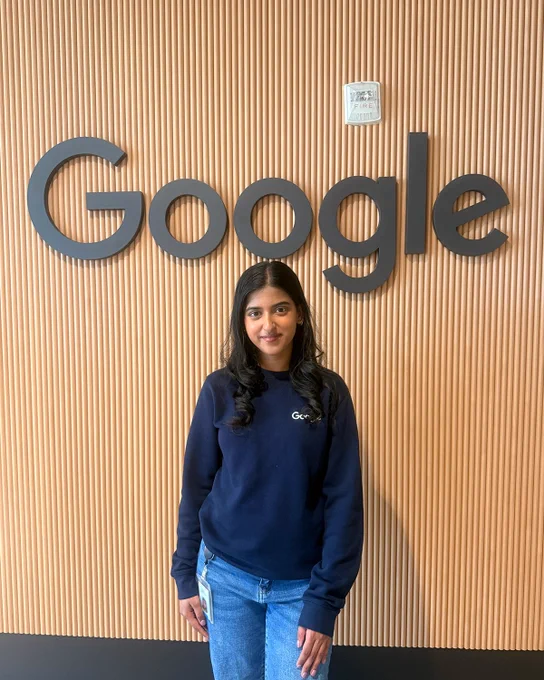 Samriddhi standing smiling in front of a light wood-paneled wall featuring a large, black "Google" logo. 

