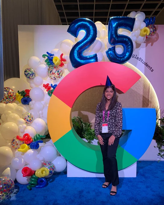 Samriddhi standing in front of a large, colorful Google "G" statue decorated with balloons and a blue "25" balloon sign. 