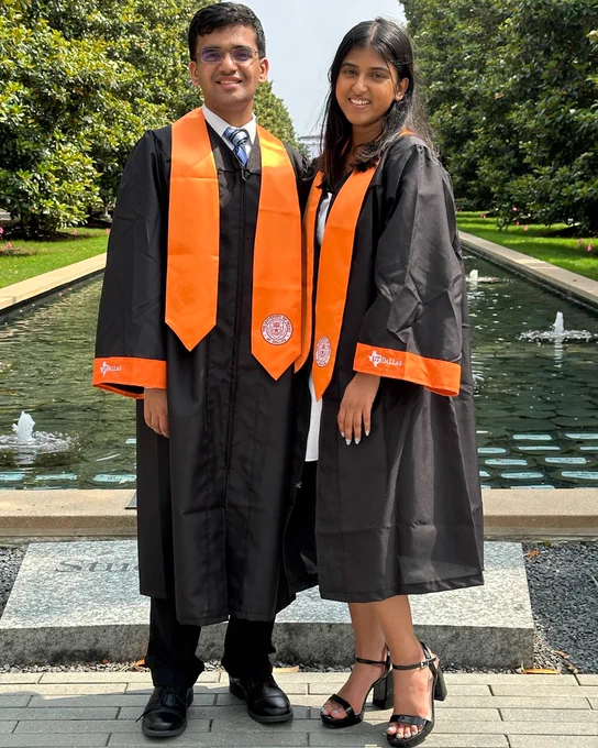 Samriddhi and a peer in black and orange graduation gowns standing outdoors in front of a pond and greenery.