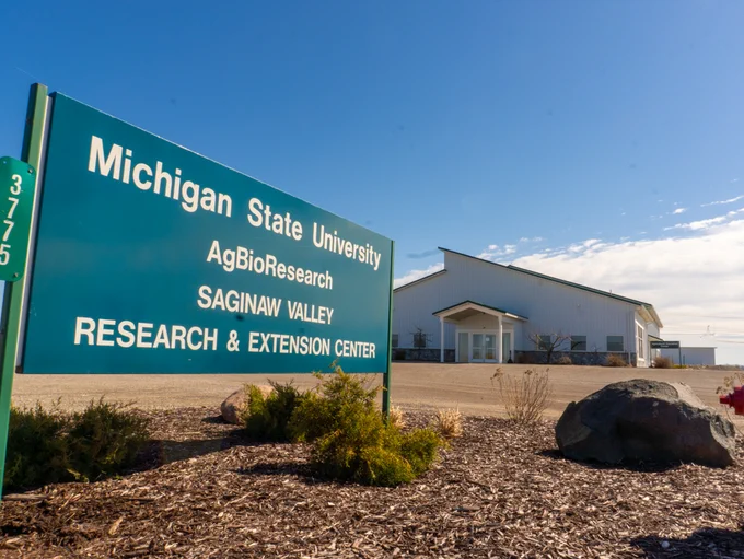 A sign reads: \"Michigan State University AgBioResearch SAGINAW VALLEY RESEARCH & EXTENSION CENTER\" with the address on a green sign. A white building stands in the background under a partly cloudy sky.
