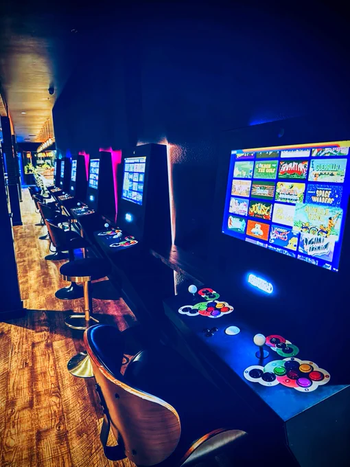 Row of Polycade arcade game machines with colorful screens and retro-style controllers in a dimly lit gaming lounge.