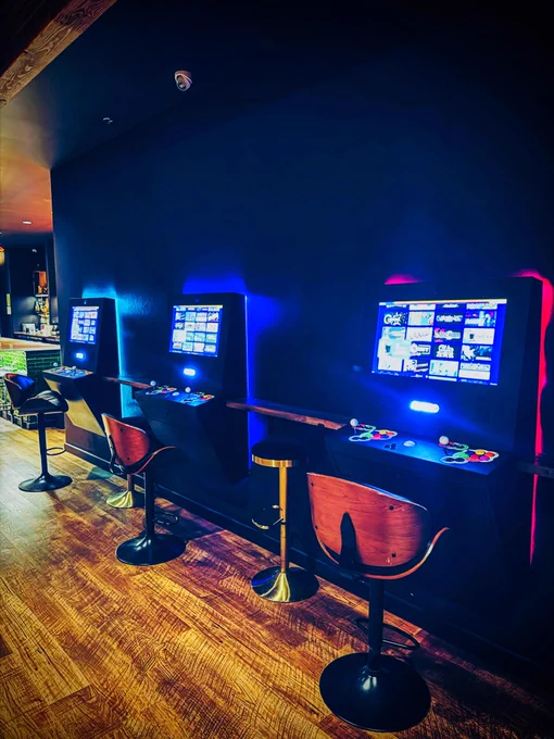 Row of illuminated Polycade arcade game stations with colorful joysticks and screens against a dark wall in a gaming lounge.