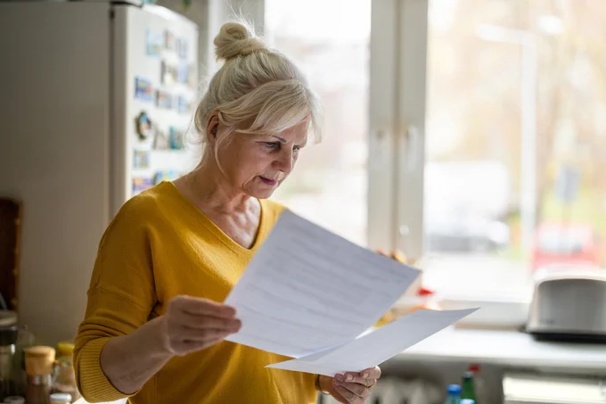 Older woman in a yellow sweater reading documents in a bright kitchen near a window.