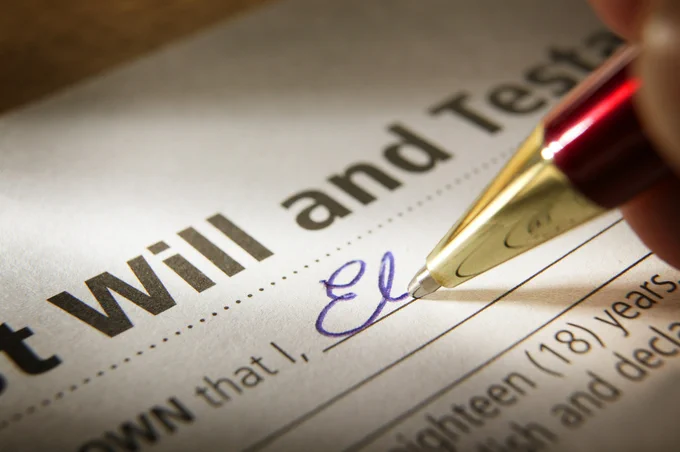 Close-up of a hand signing a last will and testament document with a gold and red pen.