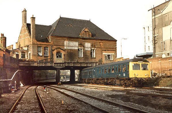 Standing on what is now the eastbound Jubilee Line looking north while a Cravens BR blue DMU runs beneath the rear elevation of the Stratford Market station building headed towards North Woolwich, on what is now the southbound DLR. All traces of platforms and stairways have been removed and the tops of the stairs bricked up save for windows. Soot on the station building's brickwork above the passenger lines is still evident from passing steam trains. Stratford High Street DLR station now stands on the right of this photo.