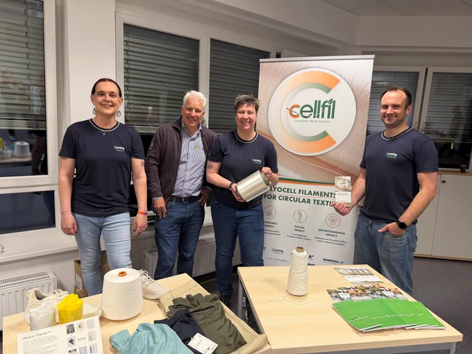 Four people standing behind a display table at an indoor event booth, posed in front of a CELLFIL roll-up banner reading “Lyocell filaments for circular textiles.” One person holds a large spool of light-colored filament yarn, while another holds a small brochure. The table in the foreground features textile-related materials, including yarn cones, fabric samples, clothing items, and informational leaflets about TENCEL™.